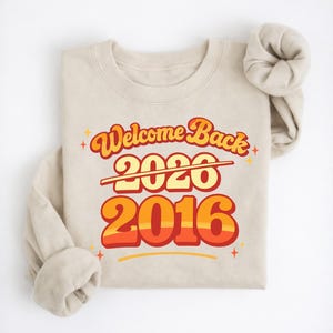 May include: Beige sweatshirt with the text "Welcome Back 2016" in retro orange and yellow font. The number 2020 is crossed out. The design includes star accents.
