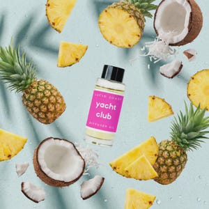 May include: A clear glass bottle of "Yacht Club" diffuser oil with a pink label, surrounded by fresh pineapple slices, whole pineapples, and coconut pieces. The bottle has a black cap and the text "Lucid Coast" above "Yacht Club".