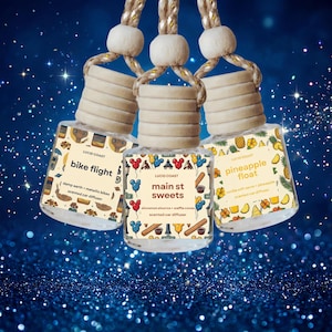 May include: Three glass car air fresheners with wooden caps and decorative labels. The labels read "bike flight", "main st sweets", and "pineapple float". Each diffuser is suspended by a beige cord with a wooden bead.