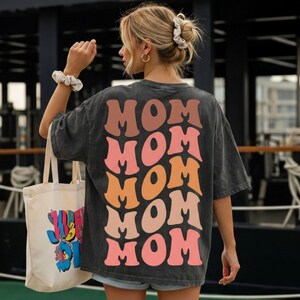 May include: Dark gray t-shirt with the word "MOM" repeated in a stacked design on the back, using shades of brown, orange, and pink. A cream-colored tote bag with a colorful graphic is visible.