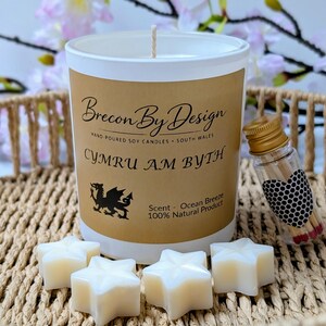 May include: A candle with a white and brown label, featuring the text "Brecon By Design" and "CYMRU AM BYTH". The candle is accompanied by star-shaped wax melts and a small glass bottle of matches. The scent is "Ocean Breeze".