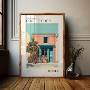 Op de afbeelding: Een ingelijste prent van een koffiehuis in Melbourne, Australië. Het gebouw is roze met een turquoise dak en deur. De tekst "COFFEE SHOP" staat bovenaan en "SINCE 1998" onderaan. Een vaas met een plant staat op een houten vloer.