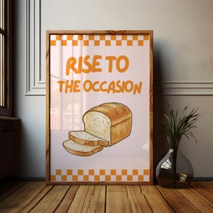 May include: Framed artwork with a loaf of bread and two slices, and the text "RISE TO THE OCCASION" in orange. The background is light pink with an orange and white checkered border. The frame is wooden.