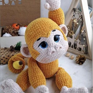 May include: A handcrafted crochet monkey toy in yellow and cream. The monkey features a detailed face with blue eyes and one arm raised. This soft plush toy is suitable for children or as a decorative item. The toy is made with soft yarn.