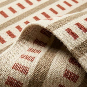 Puede incluir: Primer plano de una alfombra tejida con un patrón repetitivo de rectángulos color óxido y rayas beige sobre un fondo crema. La alfombra está doblada, revelando la textura y la construcción de la tela.