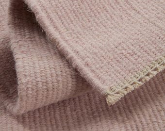 May include: A soft, pale pink woven textile featuring a dense, textured grid pattern. The fabric is folded over to reveal a light beige, fringed edge with visible thread detailing along the border, showcasing the weave and construction.