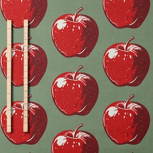 May include: A repeating pattern of red apples with white highlights and green stems on a muted green background. Two wooden rulers are vertically placed in the center of the image.