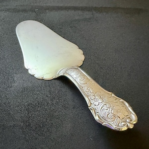 Könnte beinhalten: Silberner Tortenheber mit gewelltem Rand und aufwendigen floralen Details am Griff. Der Heber hat eine breite, flache Klinge und einen gebogenen Griff. Ein klassisches Servierutensil für Kuchen und Torten.