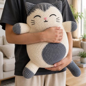 May include: A large, round, crocheted cat plush toy featuring a cream-colored body with dark gray ears, paws, tail, and a large circular patch on its belly. The cat has embroidered closed eyes, a small pink nose, and black whiskers.