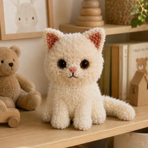 May include: A cream-colored plush cat toy made of soft, fuzzy yarn. The cat features large, dark brown eyes, a small pink stitched nose, and triangular ears with light pink inner details. It is sitting in an upright position with its tail extended.