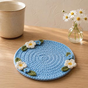 May include: A round, light blue crocheted coaster featuring a spiral stitch pattern. The coaster is decorated with small, white and yellow crocheted flowers and green crocheted leaves attached to the edges of the circular blue base.
