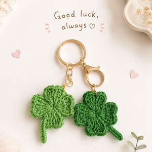 Könnte beinhalten: Zwei grüne, gehäkelte vierblättrige Kleeblatt-Schlüsselanhänger an goldfarbenen Metallringen und Karabinerverschlüssen. Über den Kleeblättern steht der Text "Good luck, always" mit einem kleinen Herzsymbol sowie kleinen rosa Herzakzenten.