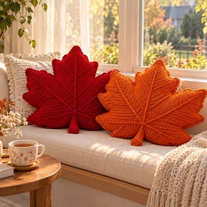 May include: Two decorative throw pillows shaped like maple leaves, one in a deep red and the other in a vibrant orange. Both pillows feature a chunky knit texture with prominent raised stitching that mimics the veins of a natural leaf.