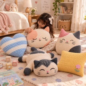 Puede incluir: Una colección de cojines de ganchillo que incluye un corazón a rayas azules y blancas, una cara de gato redonda color melocotón y blanco, un cojín cuadrado amarillo con una flor rosa y un peluche de gato tumbado de lana gris y crema.