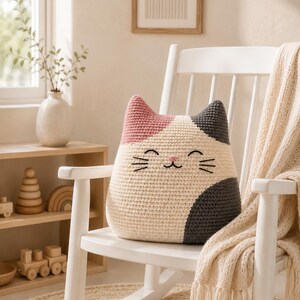 May include: A cat-shaped throw pillow featuring a crocheted texture in a tricolor pattern of cream, dusty pink, and dark gray. The pillow has embroidered facial features including closed eyes, a small pink nose, and black whiskers.