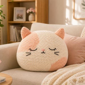 May include: A round, cream-coloured crochet cushion shaped like a cat head with peach-coloured patches on the ears and sides. The cat face features embroidered closed eyes, a small pink nose, and black whiskers stitched onto the textured yarn surface.