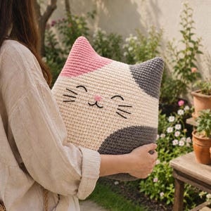 May include: A cat-shaped crochet pillow featuring a cream-colored face with a pink ear, a dark gray ear, and dark gray patches on the sides. The pillow has embroidered black eyes, a small pink nose, and black whiskers stitched onto the front.