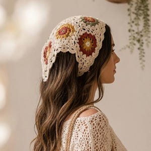 May include: A triangular crochet headscarf featuring a cream-coloured base with a repeating pattern of circular motifs in shades of mustard yellow, terracotta, and sage green. The accessory has a delicate openwork texture and scalloped edges.