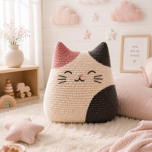 May include: A large, plush, crocheted cat-shaped pillow featuring a tricolor pattern of cream, dusty pink, and dark charcoal gray. The cat face is embroidered with black thread, showing closed eyes, a small nose, and whiskers on a cream base.