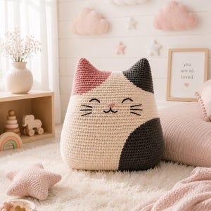 May include: A large, plush, crocheted cat-shaped pillow featuring a tricolor pattern of cream, dusty pink, and dark charcoal gray. The cat face is embroidered with black thread, showing closed eyes, a small nose, and whiskers on a cream base.