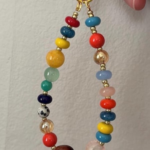 May include: A colorful beaded charm or keychain with a gold-tone clasp. The beads are a mix of round and oblong shapes in various colors, including red, blue, yellow, and pink. The charm is held by a hand.