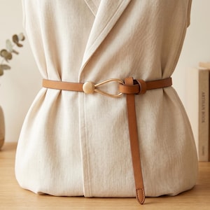 May include: A thin tan leather belt featuring a gold-toned metal buckle with a teardrop-shaped loop and a simple knot closure. The belt is worn around a sleeveless cream-coloured linen vest, creating a cinched waist effect with a long strap end.