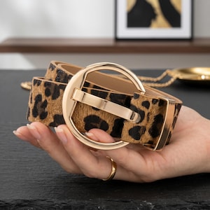 May include: A belt featuring a brown and black leopard print pattern with a textured surface. The belt is secured by a large, circular, polished gold-tone metal buckle and includes a matching gold-tone metal loop to hold the end of the strap.