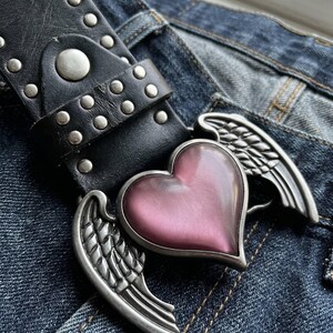 May include: Black leather belt with silver studs and a heart-shaped buckle. The buckle features a pink heart and silver wings. The belt is shown on a pair of blue denim jeans.