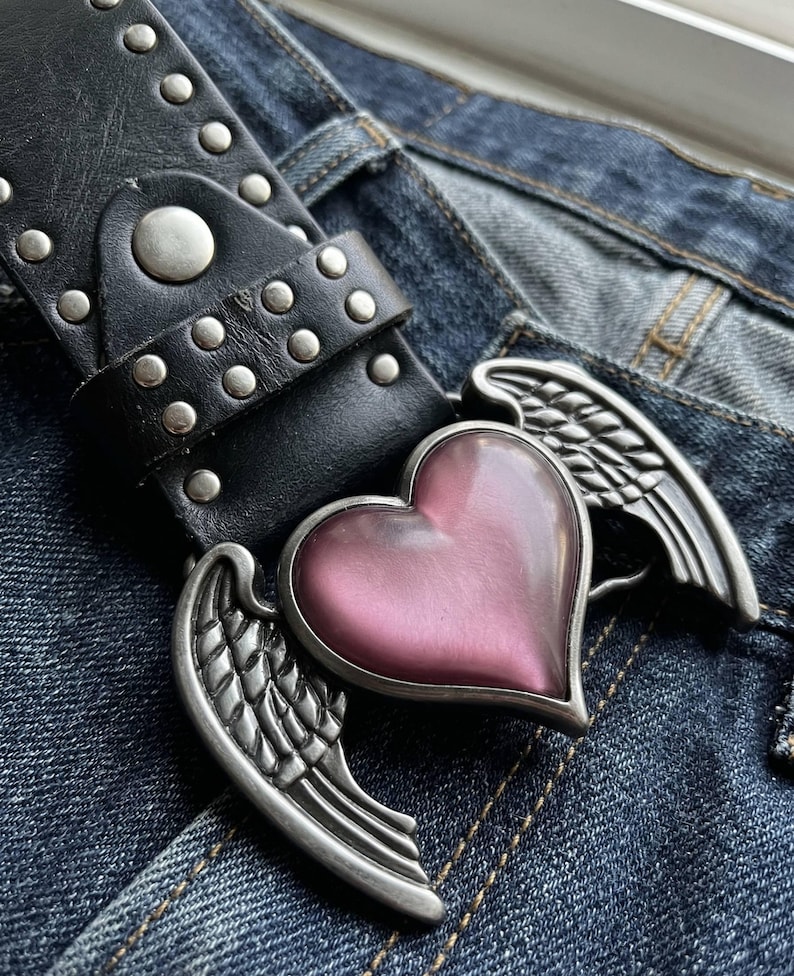 May include: Black leather belt with silver studs and a heart-shaped buckle. The buckle features a pink heart and silver wings. The belt is shown on a pair of blue denim jeans.