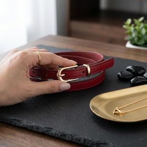 May include: A slim dark red belt featuring a polished gold-tone metal buckle and a matching gold-tone keeper loop. The belt is constructed with smooth edges and visible stitching, showcasing a classic design with a secure pin-style closure.