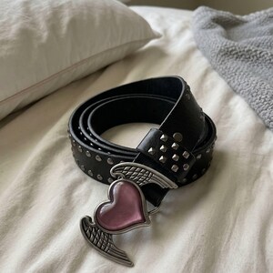 May include: Black leather belt with silver-tone studs and a heart-shaped buckle with wings. The heart is pink. The belt is rolled up on a white surface.