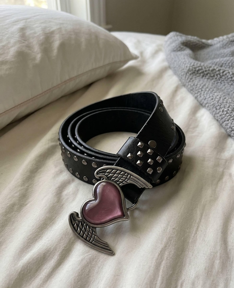 May include: Black leather belt with silver-tone studs and a heart-shaped buckle with wings. The heart is pink. The belt is rolled up on a white surface.