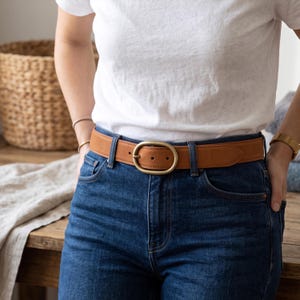May include: A tan leather belt featuring a smooth finish and a rounded, brushed brass-toned metal buckle. The belt is worn through the belt loops of dark blue denim jeans, paired with a plain white t-shirt, showcasing a classic, functional design.