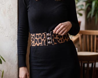 May include: A wide elastic belt featuring a tan and black leopard print pattern. The belt is secured at the center front with a silver-toned metal clasp consisting of three circular, vertically aligned interlocking hardware pieces.