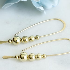 May include: A pair of gold earrings. Each earring has a curved wire design, a series of gold beads, and a flat, paddle-shaped accent. The earrings are displayed on a white marble surface.