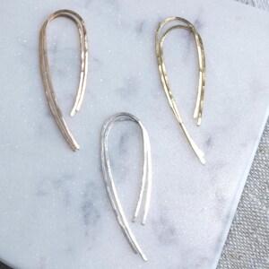 May include: Three pairs of minimalist earrings in gold, silver and rose gold. Each pair features two thin, hammered metal wires that form a simple, curved shape.