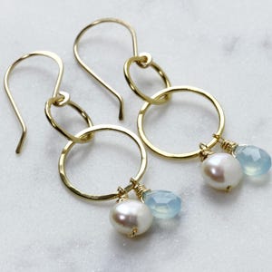 May include: Gold hoop earrings with a hammered texture, featuring a small pearl and a light blue gemstone dangling from each hoop.