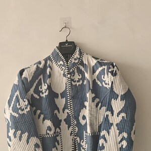 Puede incluir: Chaqueta acolchada con un estampado ikat azul y blanco. La chaqueta tiene cuello alto, mangas largas y cierre de botones. Cuelga de una percha negra.