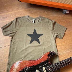 May include: Olive green t-shirt with a large black star graphic. A black and red glitter electric guitar is in the foreground. An orange amplifier with the word "ORANGE" is in the background.