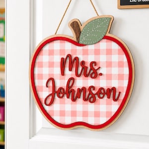 May include: A wooden apple-shaped door sign featuring a light pink and white gingham pattern. The sign has a green leaf accent and the text Mrs. Johnson in red script lettering. It hangs from a brown twine string against a white door surface.
