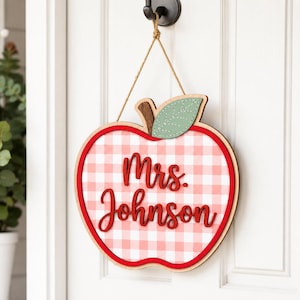 May include: A wooden apple-shaped wall sign featuring a pink and white gingham pattern. The sign displays the text Mrs. Johnson in red script lettering. A small green wooden leaf with stitched detailing is attached to the top, and a jute rope is used for hanging.