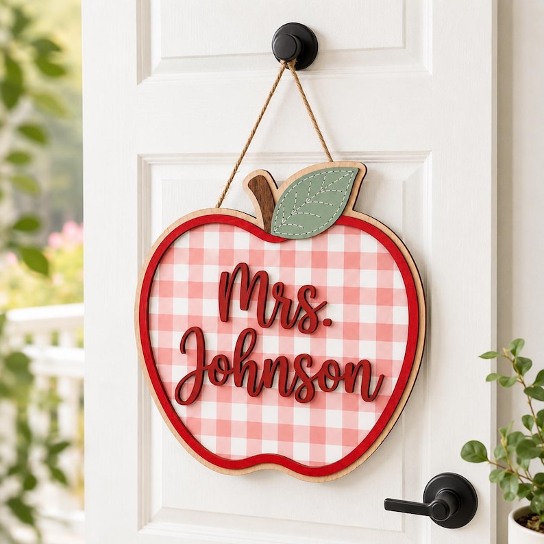 May include: A wooden apple-shaped door sign featuring a red and white gingham pattern. The sign displays the text "Mrs. Johnson" in raised red script lettering and includes a green leaf accent at the top, suspended by a thin brown twine rope.