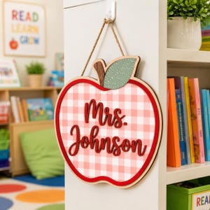 May include: A wooden apple-shaped wall sign featuring a pink and white gingham pattern. The sign displays the text Mrs. Johnson in raised red script lettering and includes a green leaf accent at the top, suspended by a brown twine rope.