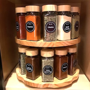 May include: A two-tiered wooden spice rack filled with glass jars of various spices. The jars have wooden lids and labels with spice names like "Cayenne," "Cilantro," and "Garlic Powder." The rack is inside a wooden cabinet.