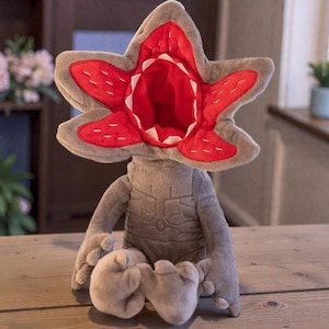 Pode incluir: Uma caneca de cerâmica branca com uma alça preta e o texto "But First, Coffee" em negrito preto. A caneca está sobre um fundo desfocado, sugerindo um ambiente acolhedor.