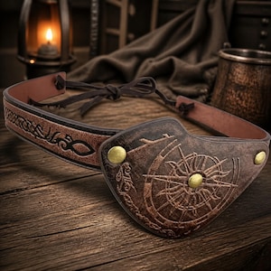 May include: A brown leather eye patch featuring an embossed compass rose design and three brass-colored metal studs. The eye patch is attached to a thin leather strap with intricate knotwork patterns and dark brown leather ties for adjustment.