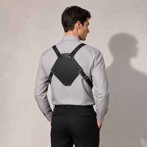 May include: A black leather-like chest harness featuring a pentagonal back panel with silver-toned metal rivets and adjustable black elastic straps. The harness is worn over a light gray long-sleeved button-down shirt and black trousers.