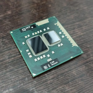 Może przedstawiać: Zielony procesor komputera z dwoma ciemnymi prostokątnymi elementami i licznymi małymi srebrnymi pinami. Procesor ma tekst "INTEL" i inne kody. Przedmiot znajduje się na ciemnej drewnianej powierzchni.