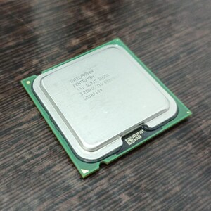 Może przedstawiać: Srebrny procesor Intel Pentium z zieloną podstawą płytki drukowanej. Procesor ma nadrukowany tekst na swojej powierzchni, w tym logo Intel i informacje o modelu. Procesor ma kształt kwadratu i leży na drewnianej powierzchni.