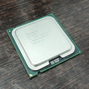 Może przedstawiać: Srebrny procesor Intel z tekstem na górnej powierzchni, umieszczony na zielonej płytce drukowanej. Procesor ma kształt kwadratu i leży na ciemnej drewnianej powierzchni. Tekst na procesorze jest częściowo widoczny.